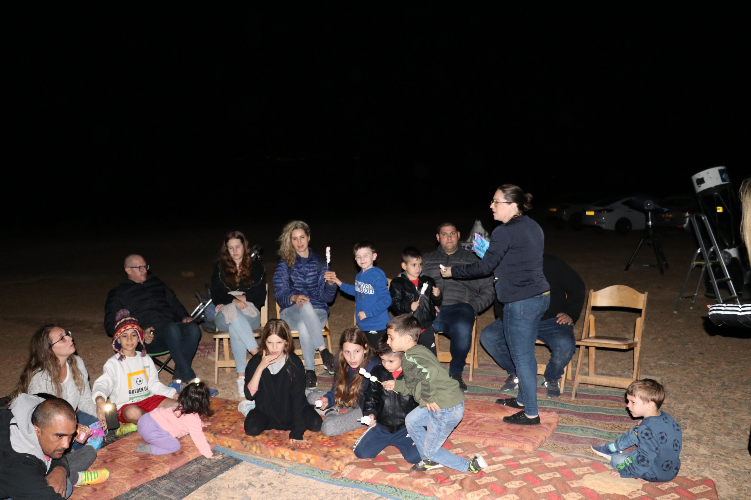 קבוצת לקוחות יושבים על שמיכות מעוצבות מסביב למדורה בלילה. הם קולים מרשמלו על מקלות. הסביבה המדברית מוארת במעומעם, כשהקבוצה נראית מאורסת ונהנית מהפעילות.