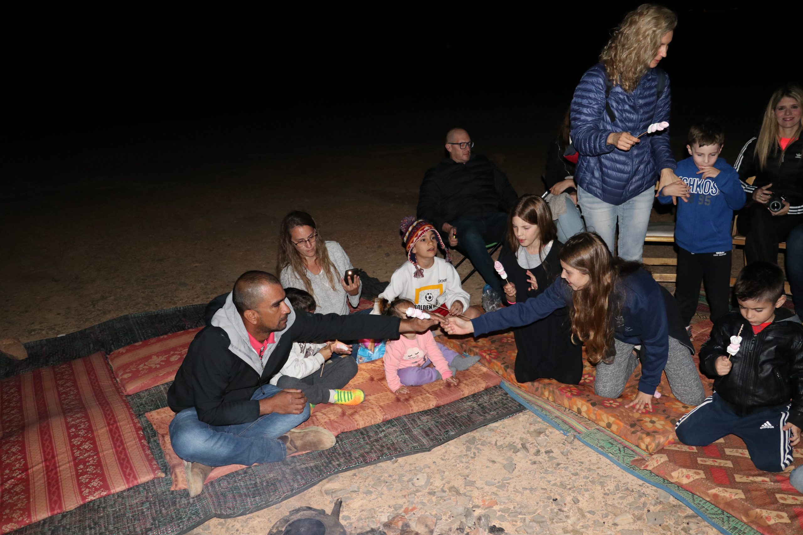 קבוצת מבוגרים וילדים יושבים על מחצלות מסביב למדורה בלילה. הם אוכלים מרשמלו על מקלות. הסביבה המדברית מוארת במעומעם, כשהקבוצה נראית מאורסת ונהנית מהפעילות.